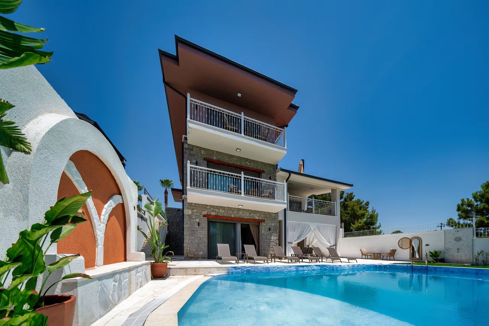 The villa's pool terrace at midday, with loungers set along the water and the stone façade behind